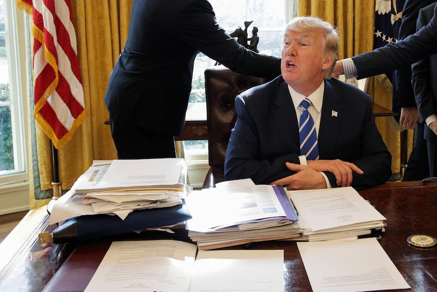 Donald Trump sits at his desk in the Oval Office. Papers are scattered all over it, and a black zipped lockbag with the key in
