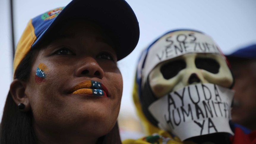 A woman wearing a cap and with face paint of the Venezuelan flag stands next a skull mask with Venezuelan words written on it.
