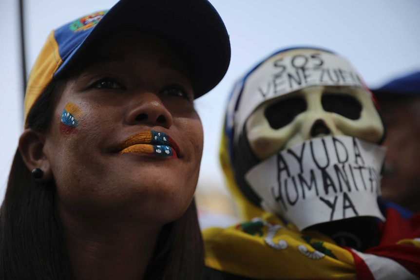 A woman wearing a cap and with face paint of the Venezuelan flag stands next a skull mask with Venezuelan words written on it.