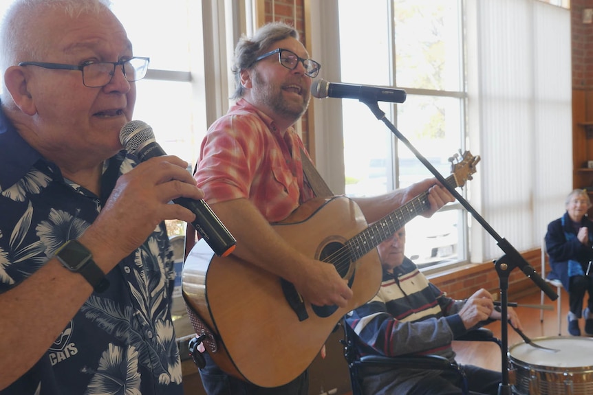 An elderly man with microphone sings accompanied by a guitarist.