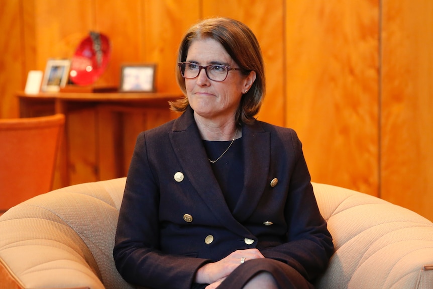 A middle-aged woman in a purple outfit and weading glasses sits on a plush chair with her hands in her lap.