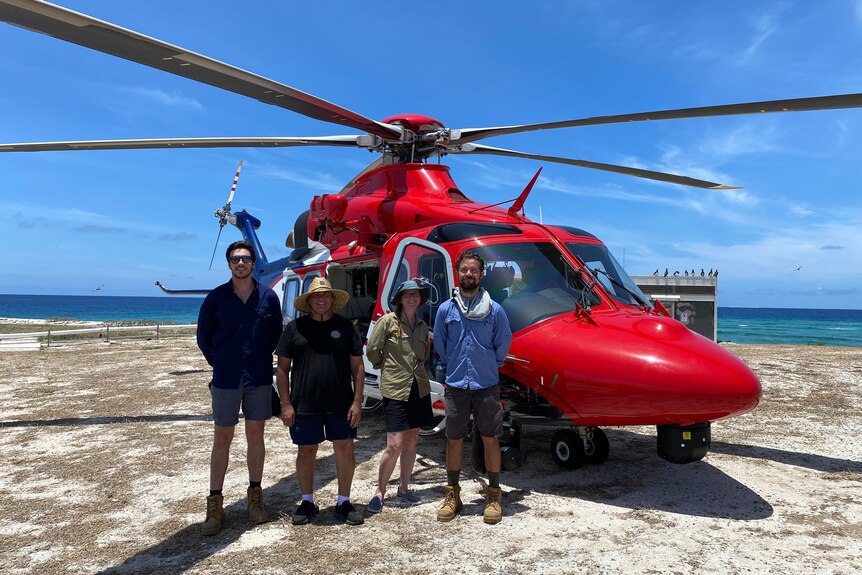 A woman and three men stand in front of a red helicopter near the ocean