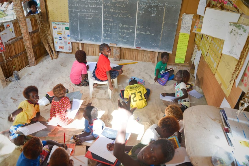 A group of smiling children learn in a school, sitting in the sand.