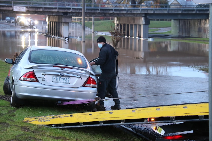A man wearing a mask checks a car abandoned near a flooded creek.