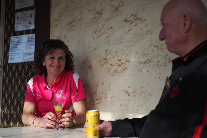 A middle aged lady with tight curly brown hair and a pink t shirt smiles at the camera while sitting at a table.