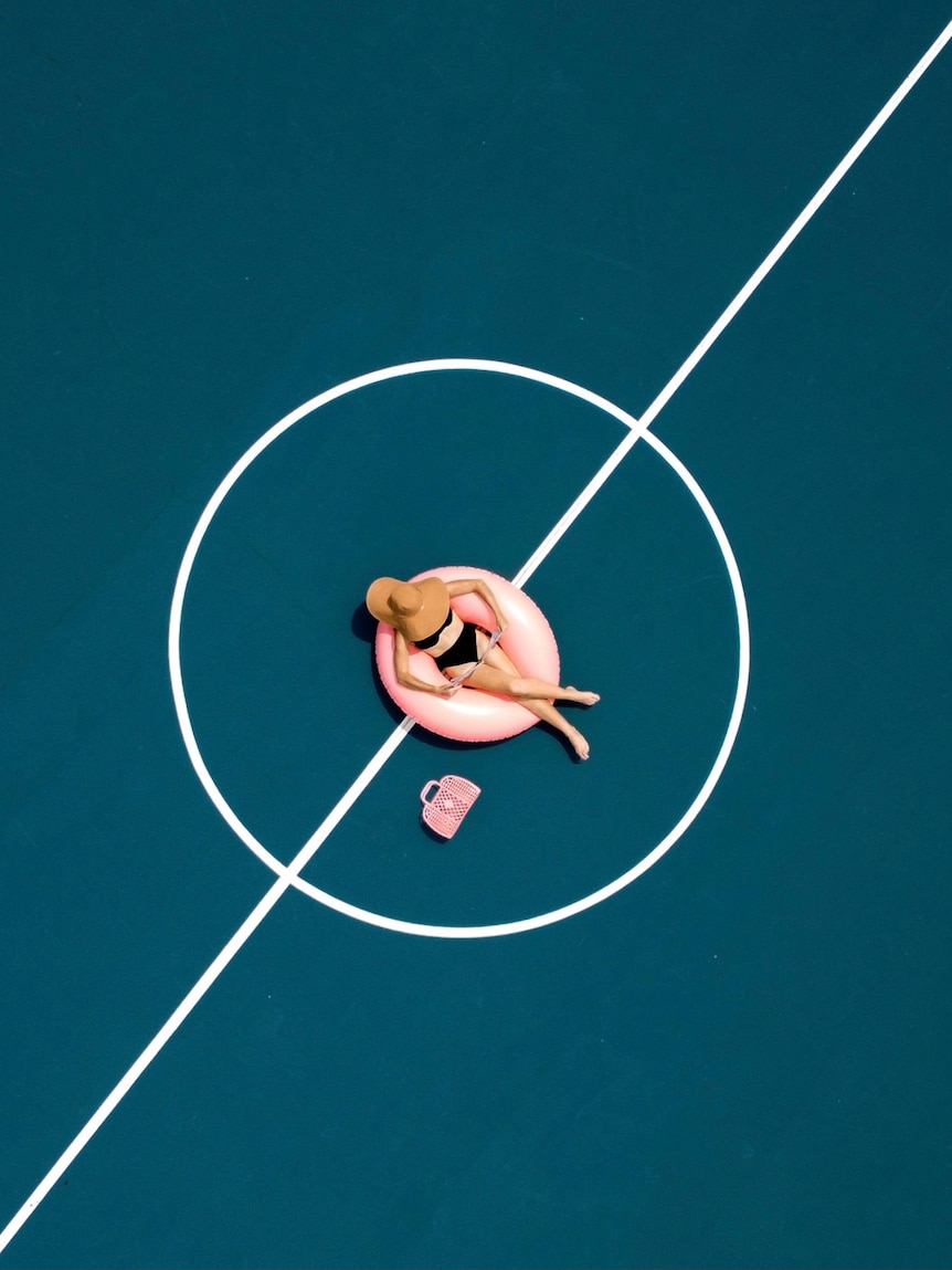 Photographed from above, a woman in a swimming costume sits in a pool floaty in the middle of a blue basketball court