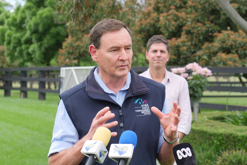 A man gestures while speaking in front of media microphones in a tree lined paddock