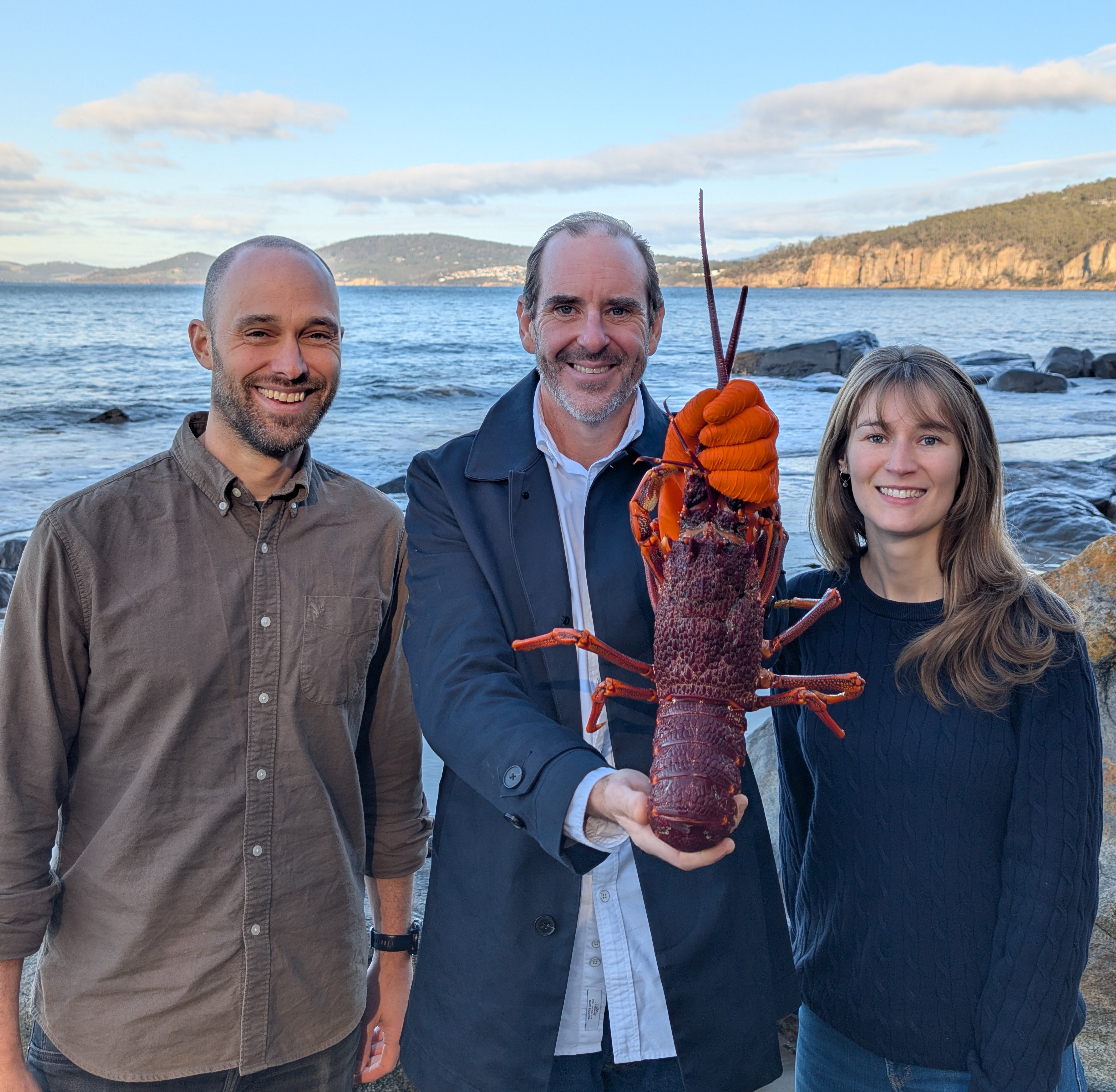 Two men and a woman smile while holding a lobster next to a body of water.