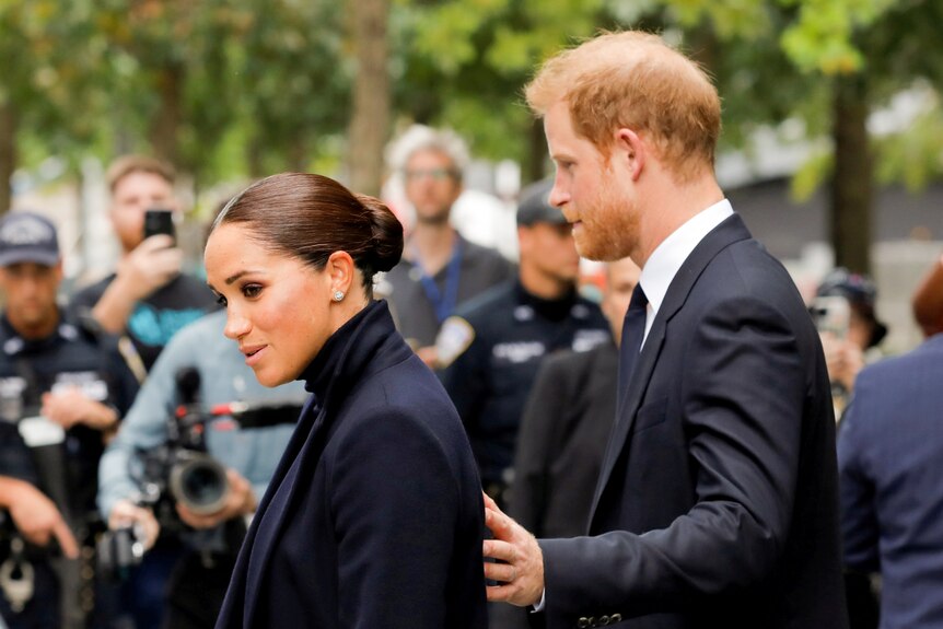 Harry puts his hand on Meghan's back in front of a media scrum.