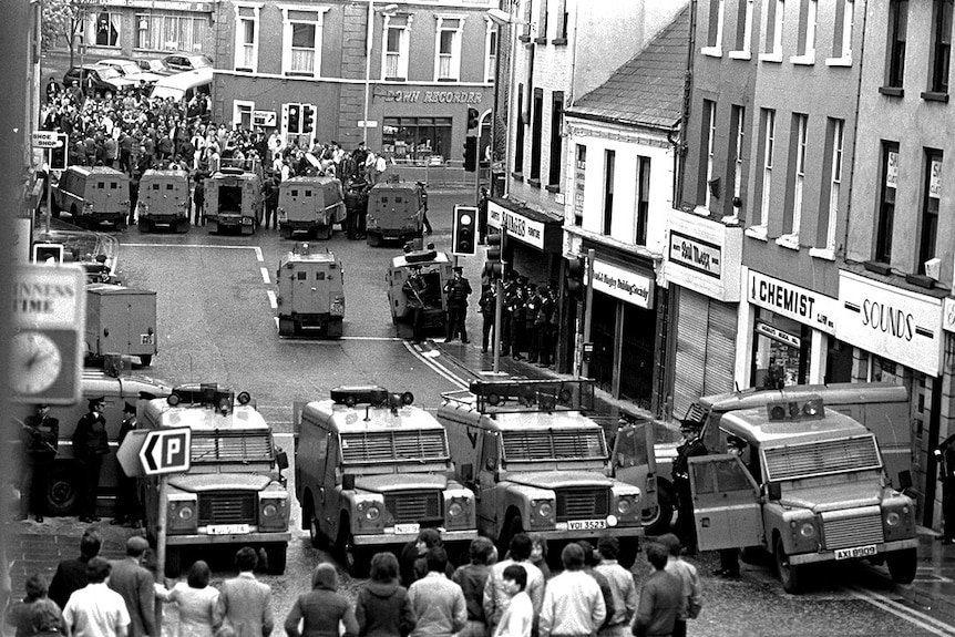 From a high angle, you look down at a street where a rows of Land Rovers separate warring groups.