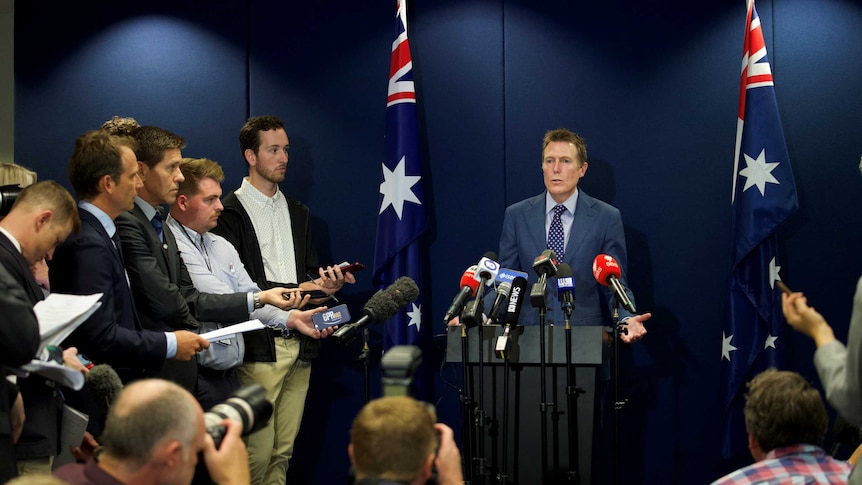 A man at a podium with microphones surrounded by journalists