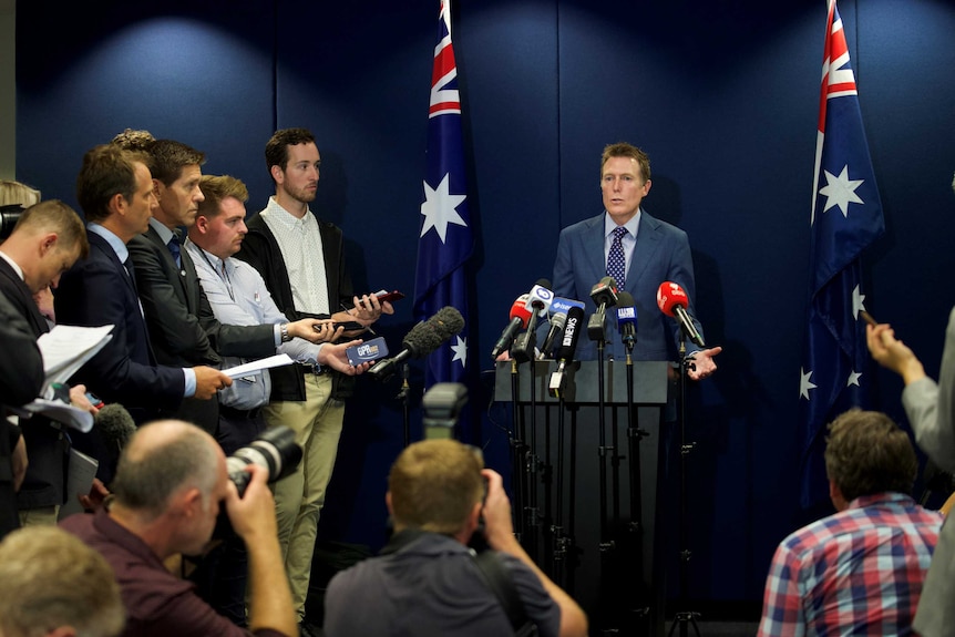 A man at a podium with microphones surrounded by journalists