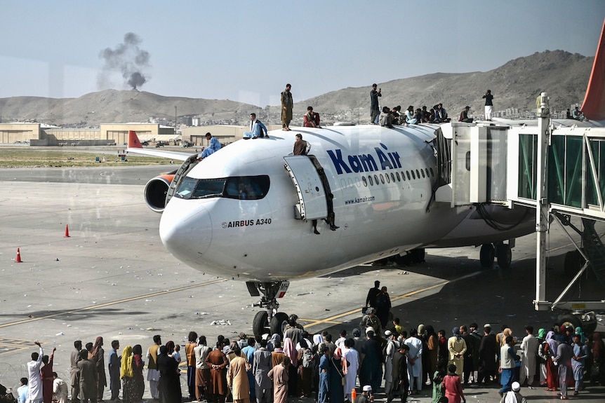 People stand on top of a commercial plane at an airport. A crowd of people stand in front of it on the tarmac