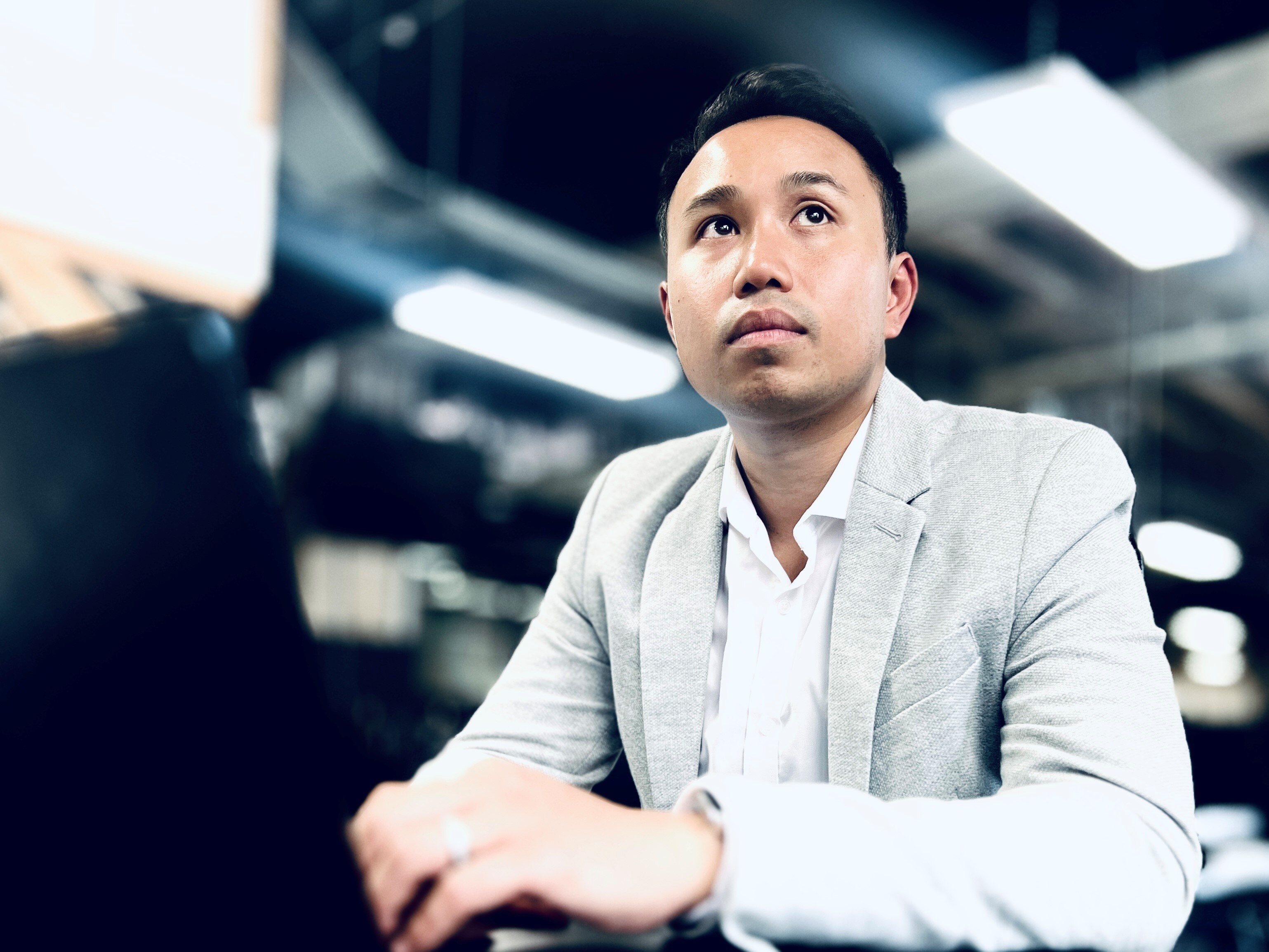 A man wearing a grey blazer and white business shirt sits at a desk in front of a laptop looking into the distance.