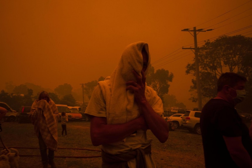A photograph of a bushfire-affected community. The sky is red with smoke. A man wears a towel over his face.