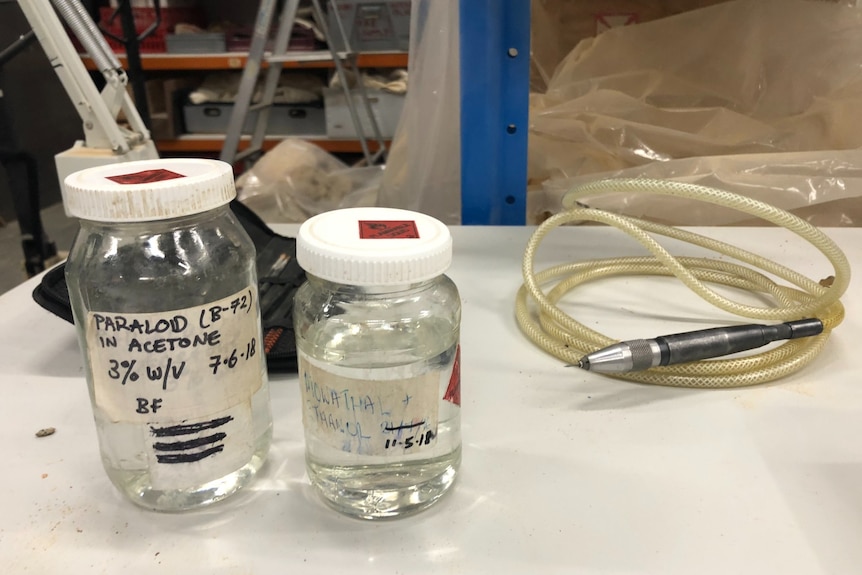 Two glass jars with a white lid containing clear liquid in front of a drill and air hose.