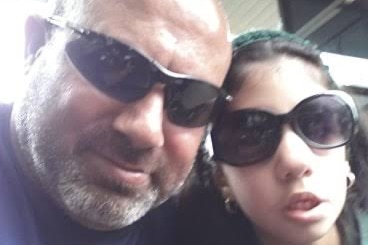 A man wearing dark sunglasses and a blue t-shirt takes a photo with his daughter who is also wearing sunglasses