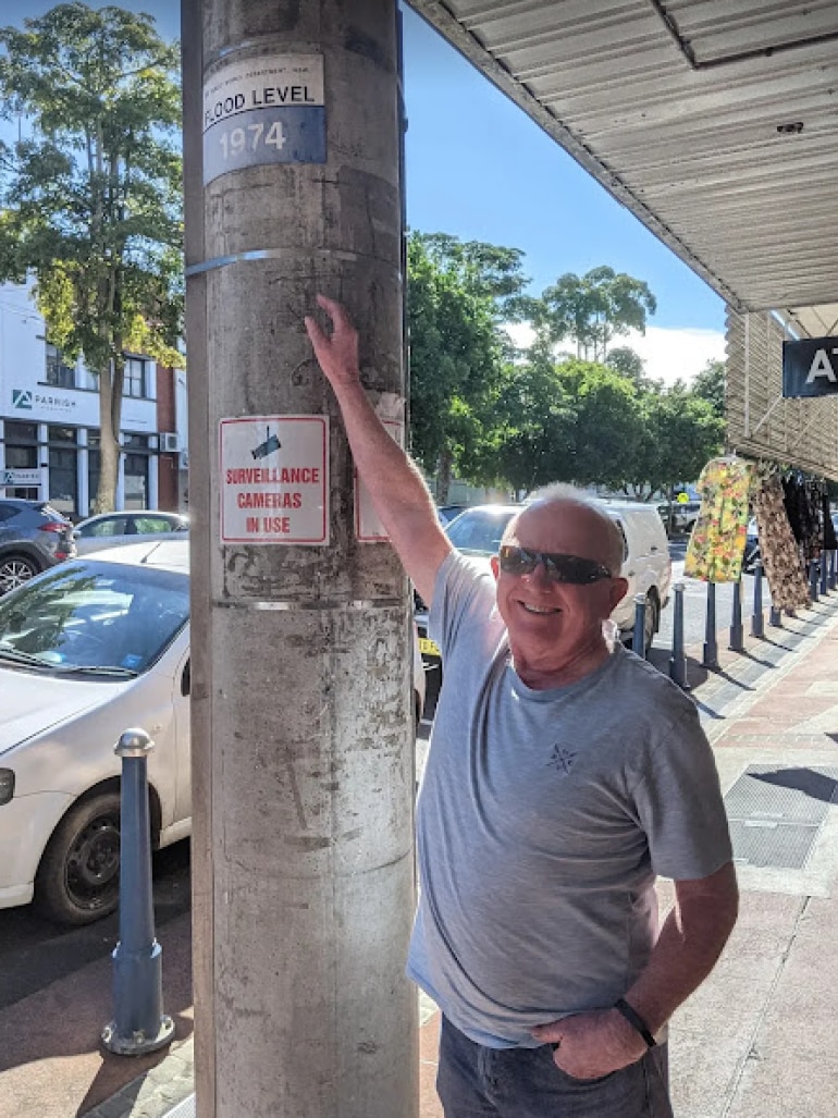 Man stands next to telegraph pole on CBD footpath, reaching his arm high above his head to reach mark on the pole