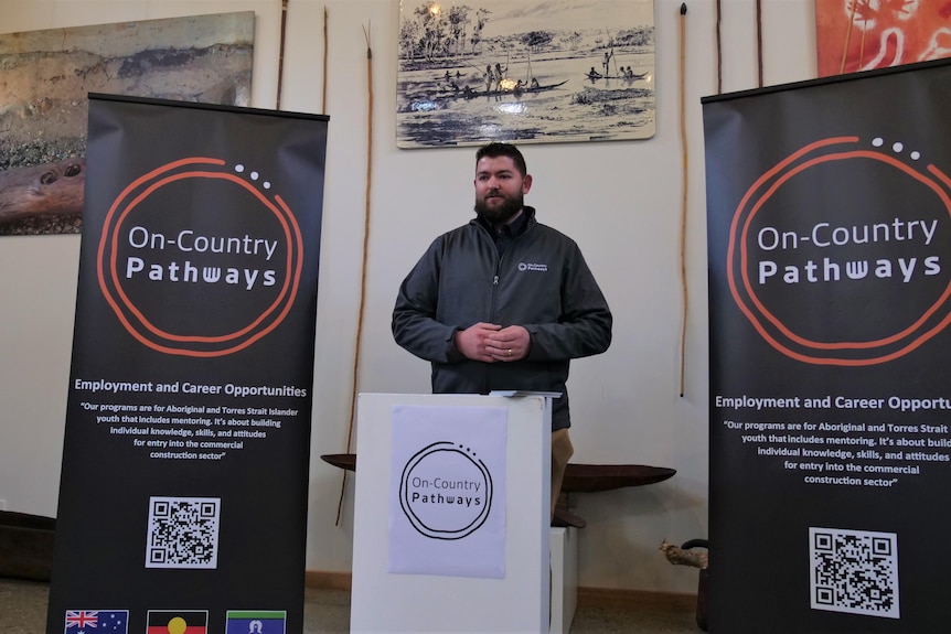 A man in a grey jacket stands at a podium in front of on-country pathways