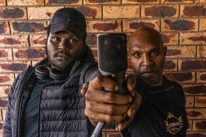 Father and son stand back against a brick wall holding a microphone