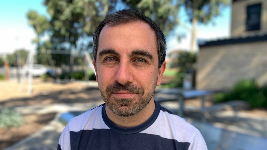 Close up of a man wearing a white and navy striped t-shirt, with trees in the background.