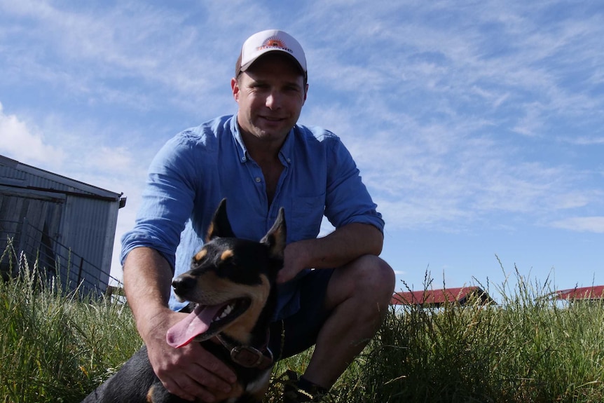 Man wearing a cap crouching behind a kelpie that's sitting down with sheds and a big blue sky behind them