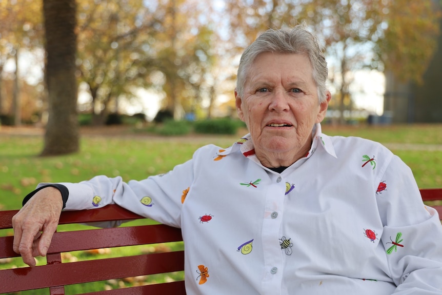 A woman with short silver hair sits on a park bench in a white shirt embroidered with insects
