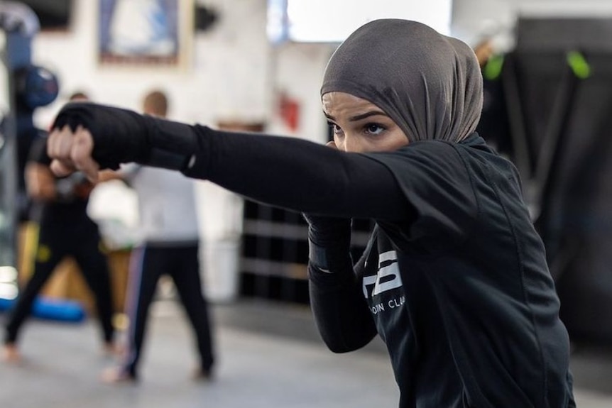A woman wearing black clothes and a grey hijab performs a boxing punch