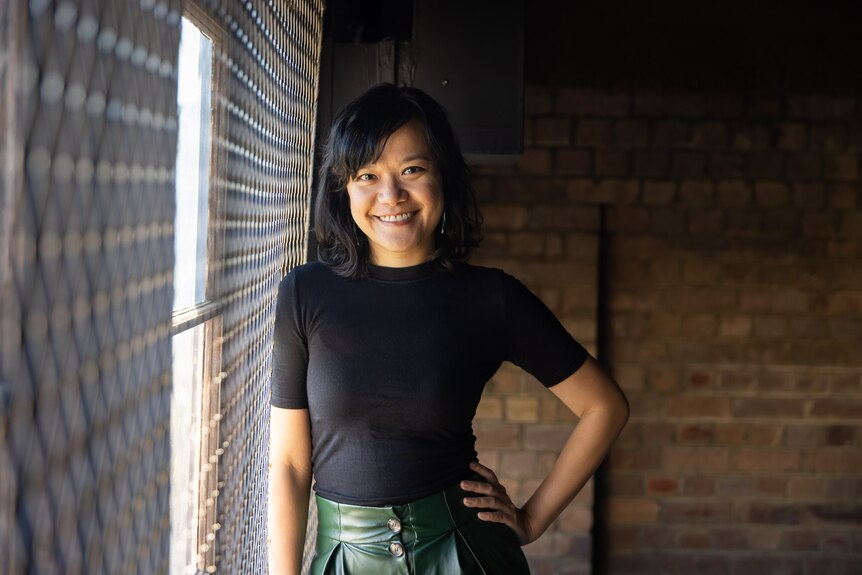 A smiling 30-something Asian Australian woman in a black shirt and green trousers stands with her hand on her hip