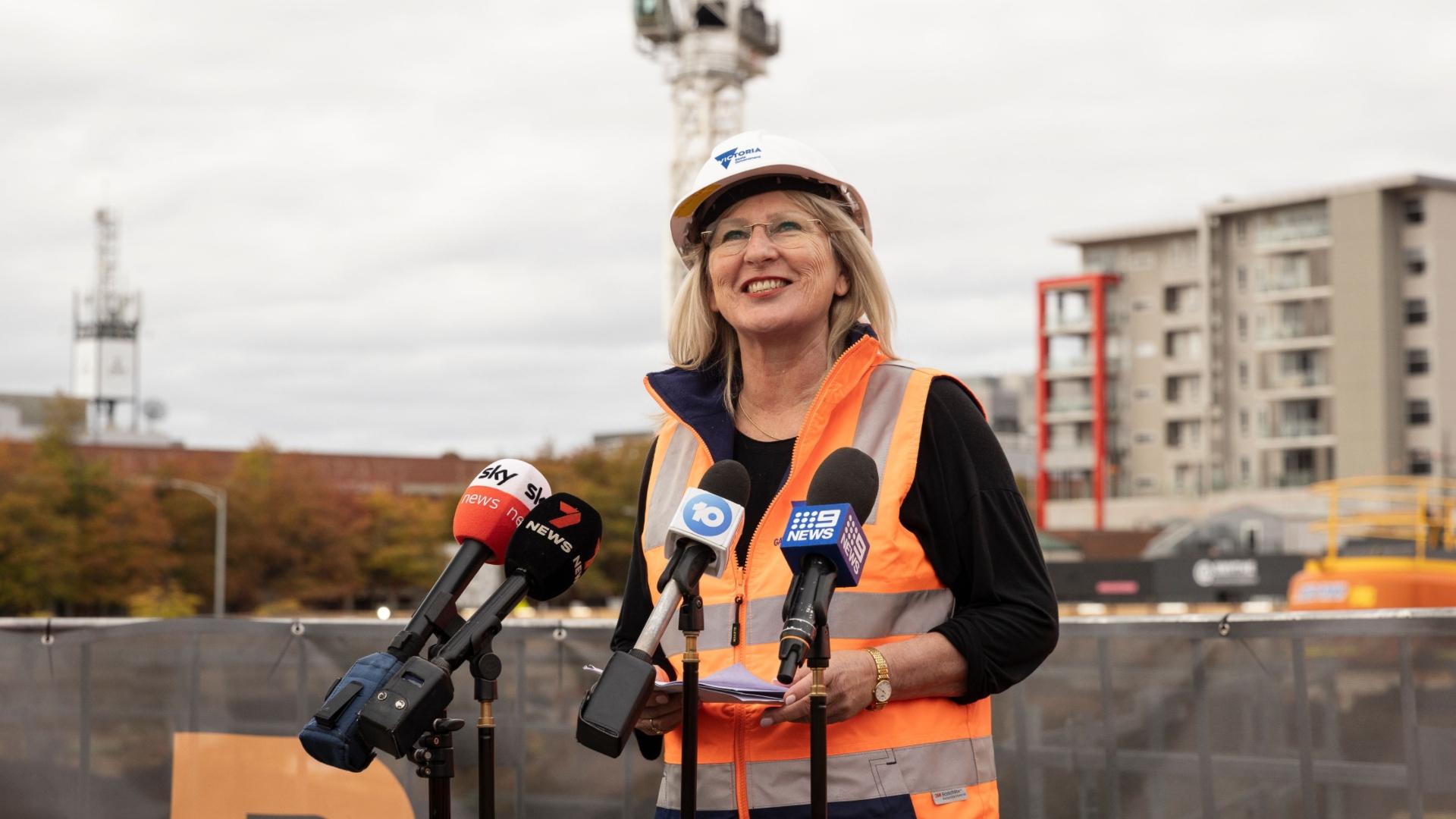 Женщина в яркой одежде и каске говорит в микрофоны перед строительной площадкой.
