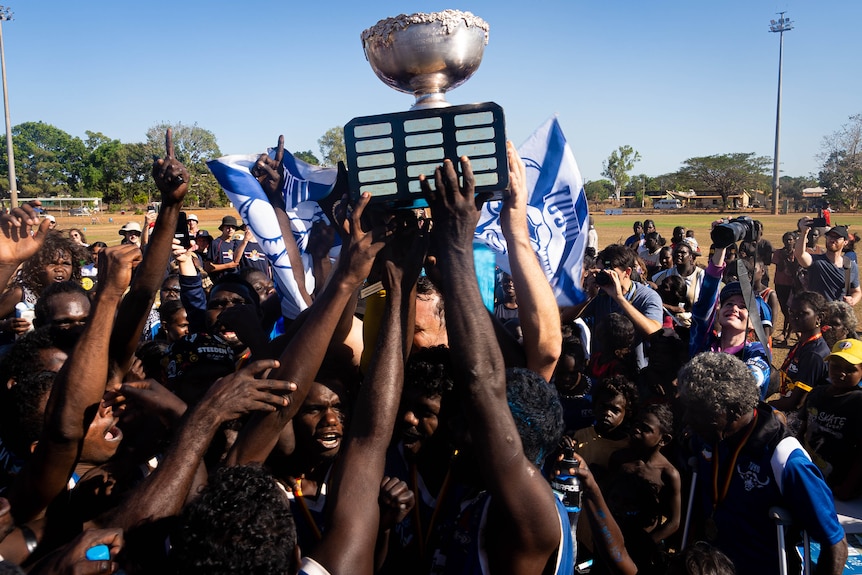 A large group of people hold up a large silver trophy. Someone holds a blue flag in the background 