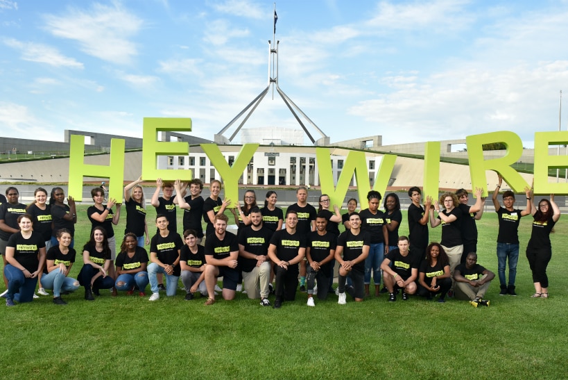 A large group of young people wearing Heywire T-shirts and holding giant letters that spell Heywire in front of Parliament House