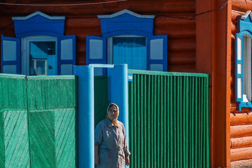 A woman stands wearing a head scarf against a backdrop of bright green, blue and orange buildings