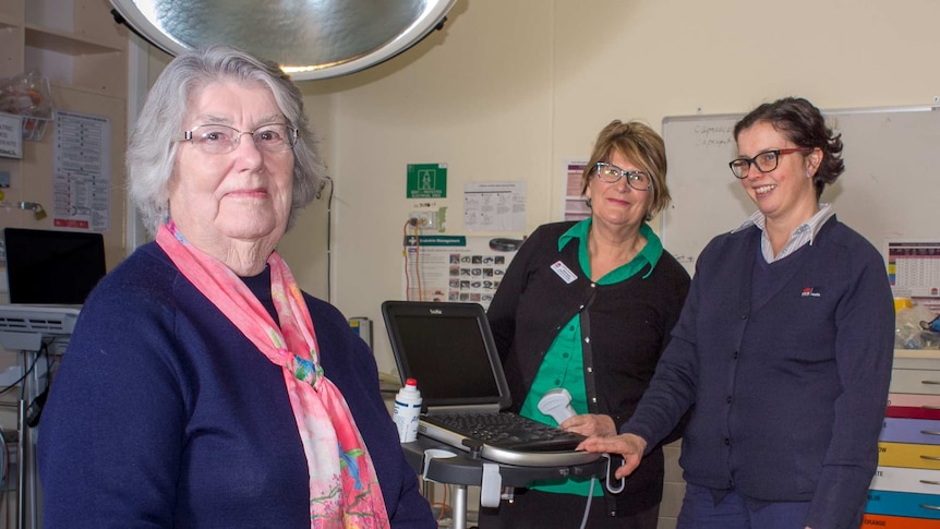 An older woman standing in a small hospital room with two women standing behind looking at an ultrasound machine