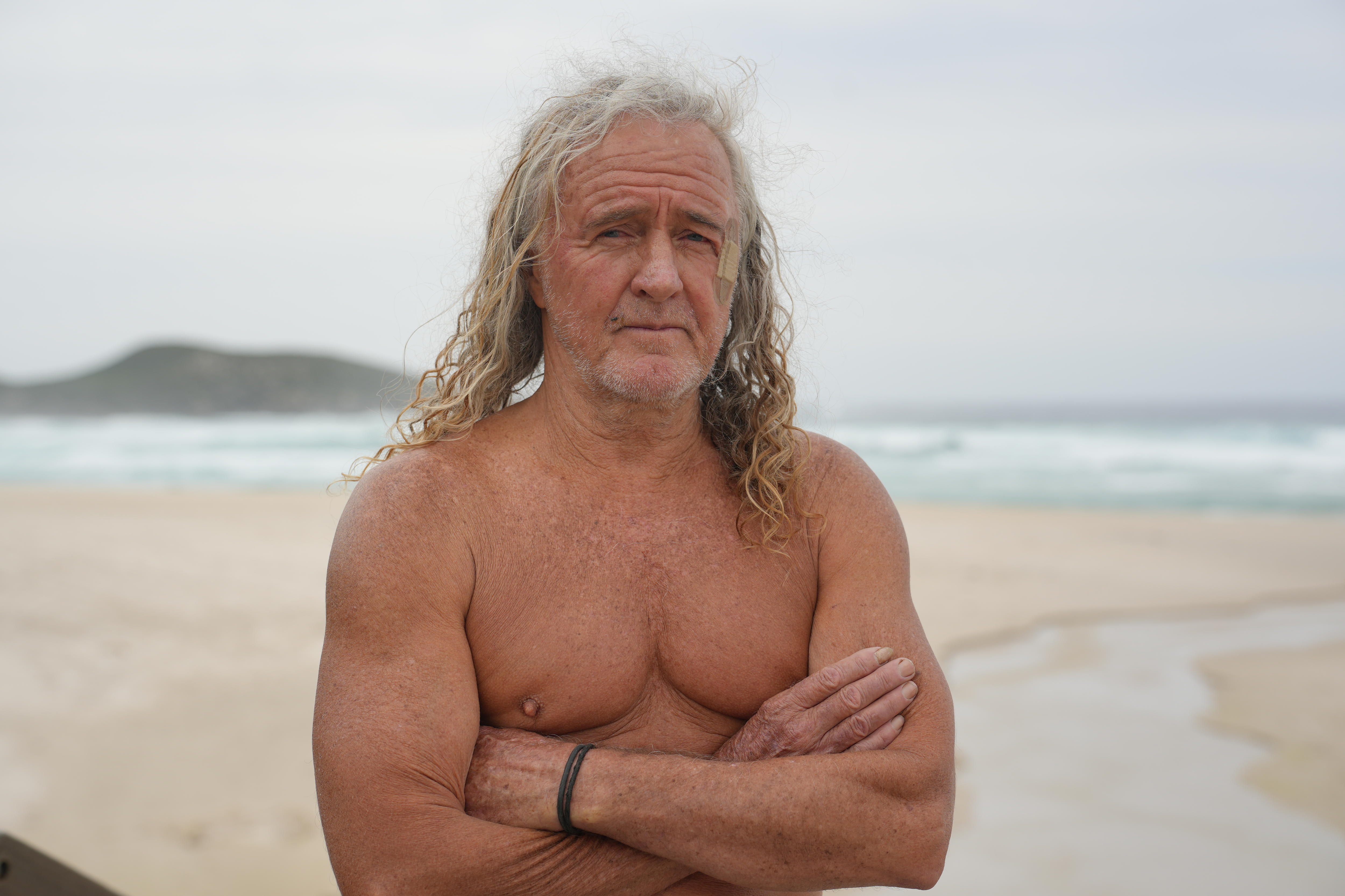 A shirtless man with long grey hair on a beach.  