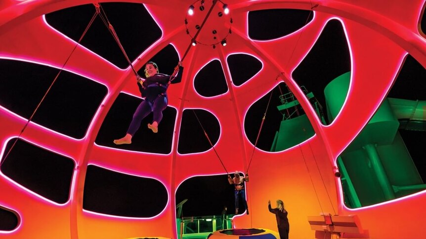 Inside a large red dome, a woman wearing black clothes is bouncing on a trampoline while suspended by bungee cords.