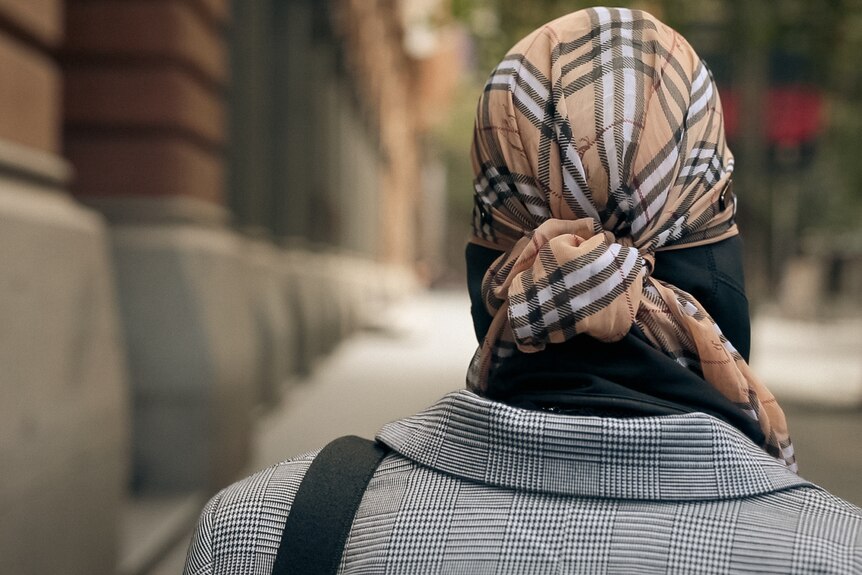 The back of a person's head, as they walk past a sandstone building wearing a Burberry scarf on their head.