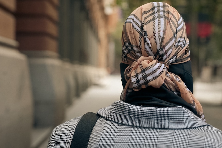 The back of a person's head, as they walk past a sandstone building wearing a Burberry scarf on their head.