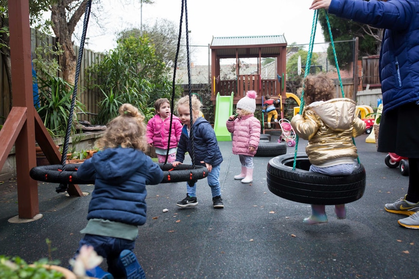 Children play on swings and others climb on a cubby house in the background.