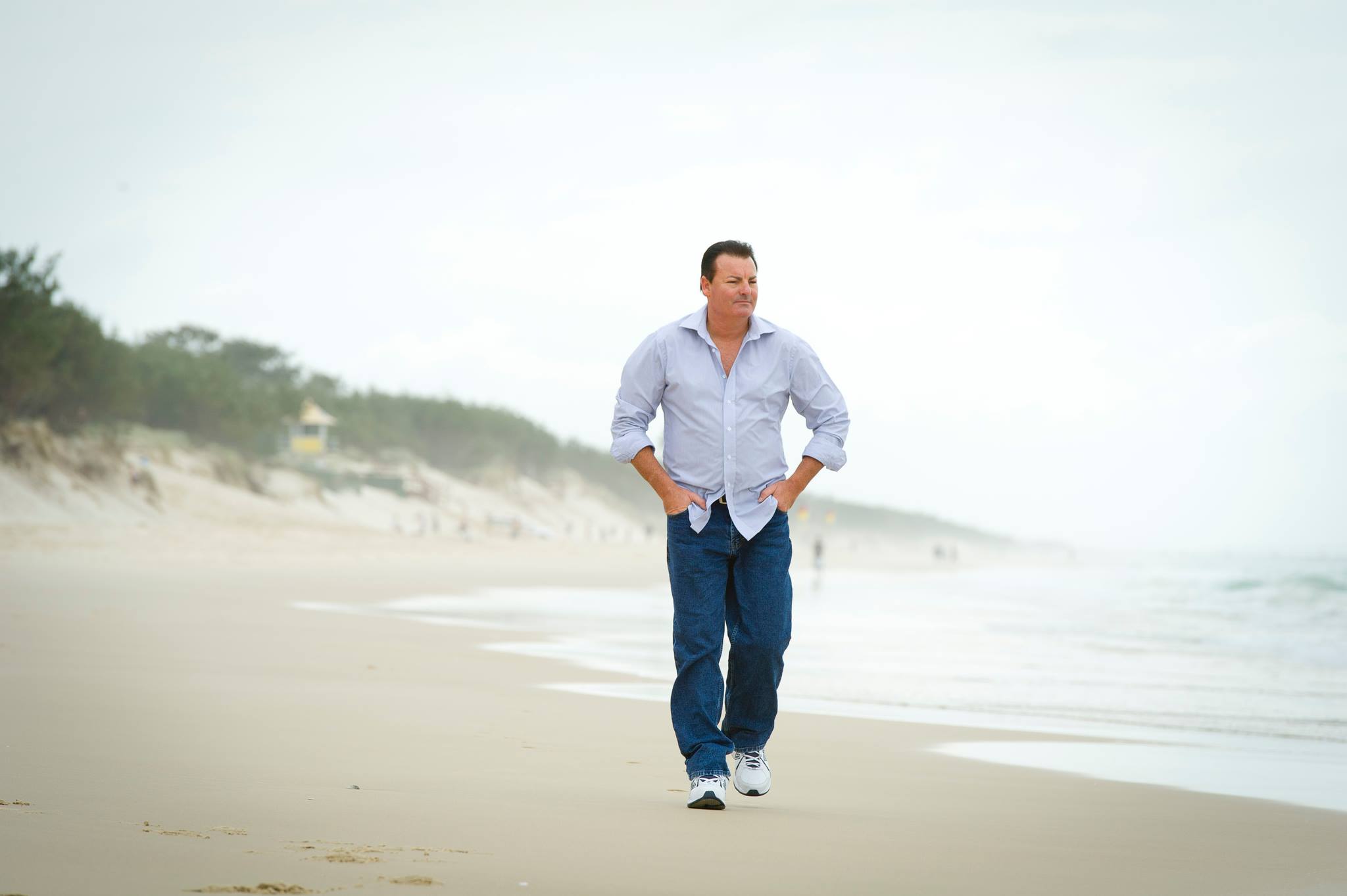 A man wearing jeans and a blue shirt walks on a beach.