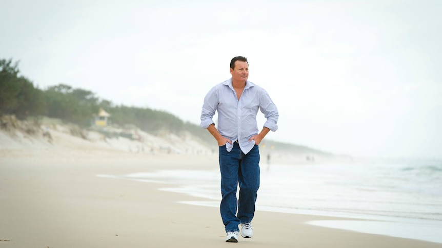 A man wearing jeans and a blue shirt walks on a beach.