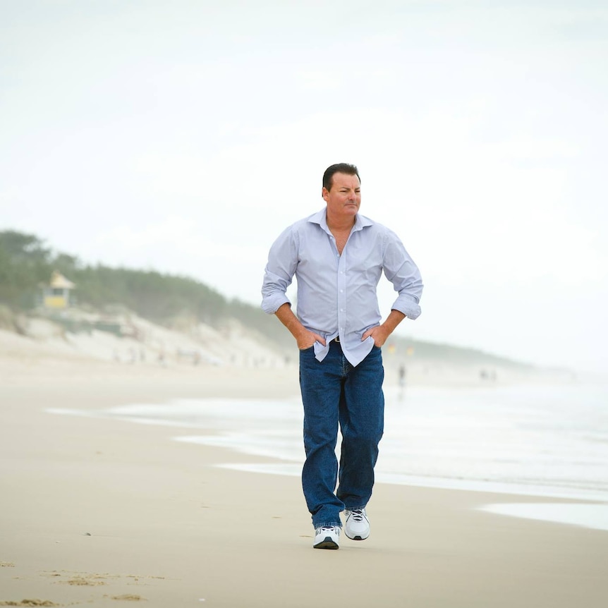 A man wearing jeans and a blue shirt walks on a beach.