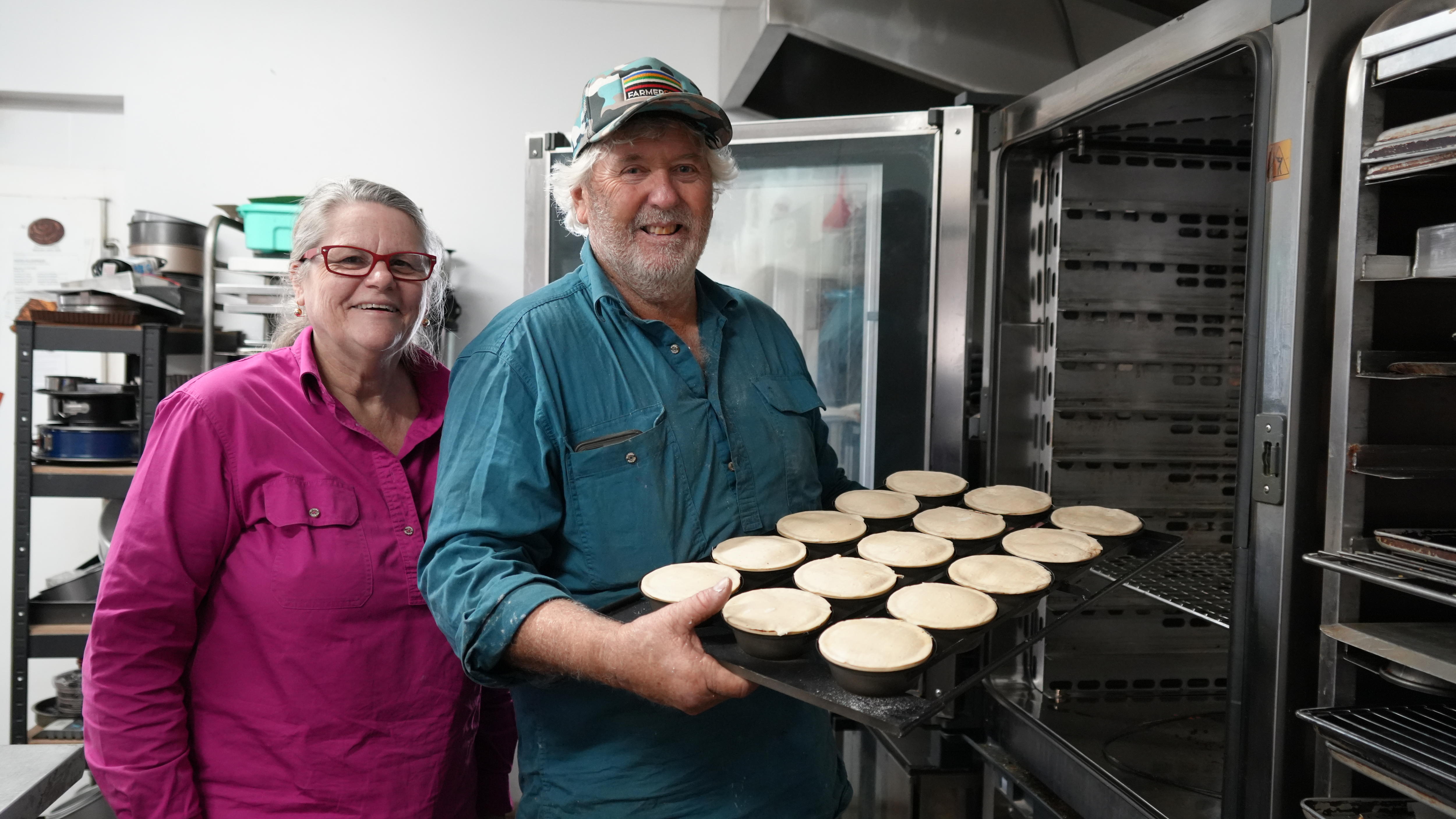 A middle aged man and woman smiling holding a tray of uncooked pies next to an oven.