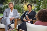 Prince Harry and Megan sit on armchairs, looking towards Oprah Winfrey, whose back is to the camera.