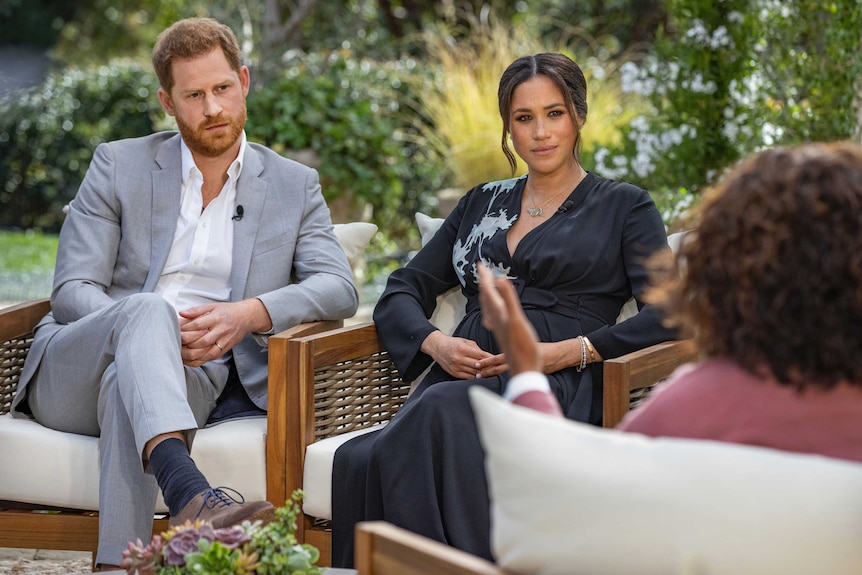 Prince Harry and Megan sit on armchairs, looking towards Oprah Winfrey, whose back is to the camera.