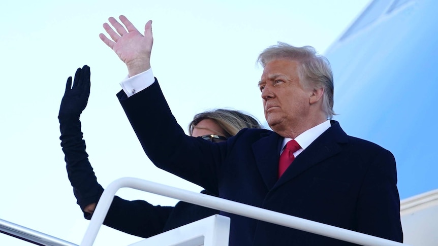 A man in a suit with a red tie waves next to a woman waving on steps