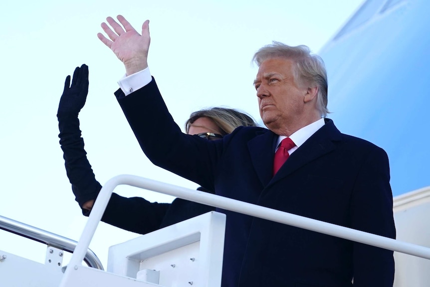 A man in a suit with a red tie waves next to a woman waving on steps