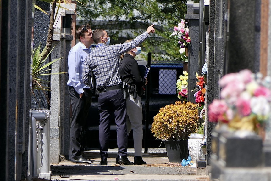 Three police officers stand in the cemetery, with one officer pointing at a grave