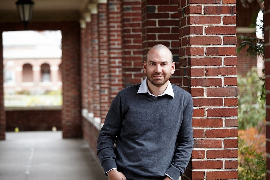 a bald man leaning against a brick column. 