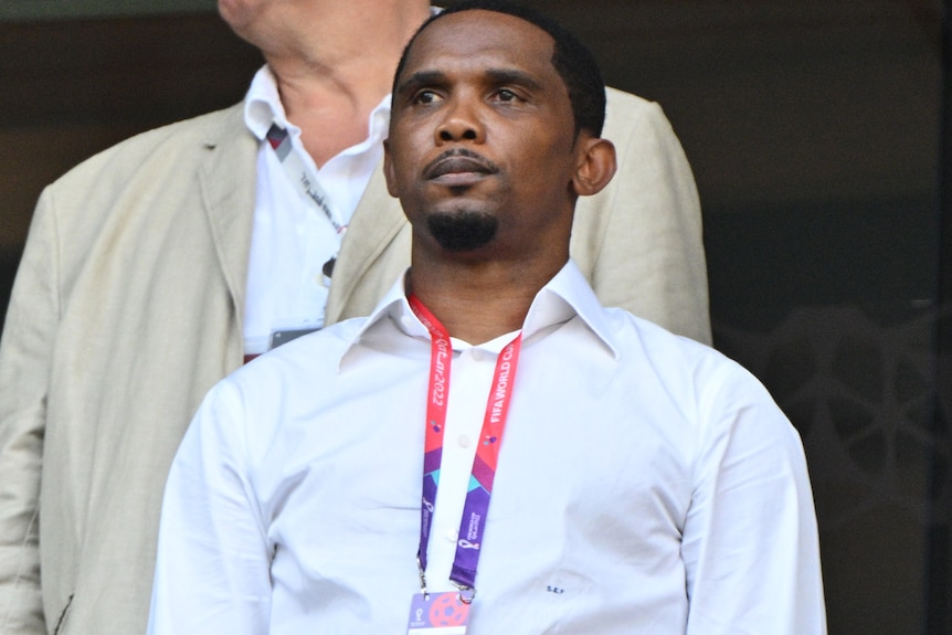 A man in a white shirt stands, he has a lanyard around his neck.